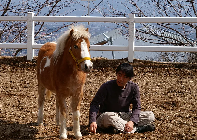 長野の乗馬クラブの馬場で、ポニーの横に地べたに座っている西達郎
